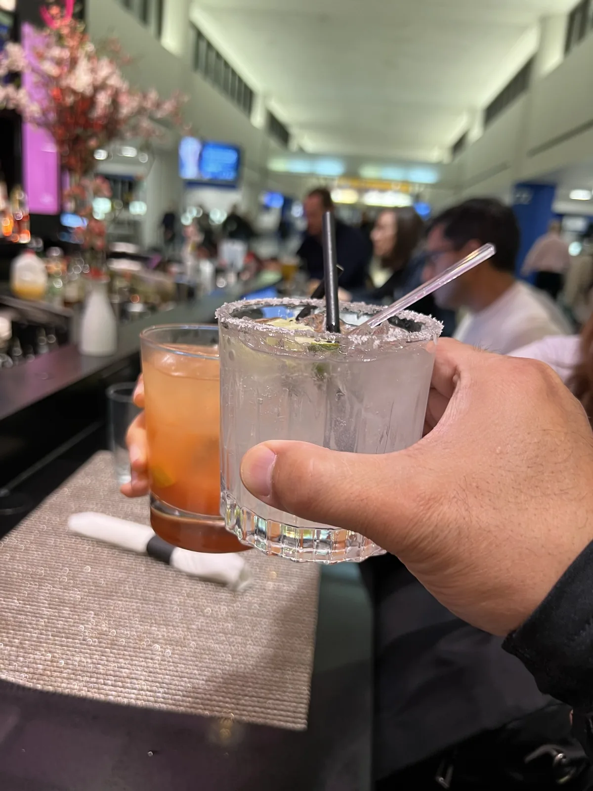 Two cocktails at an airport bar, with the terminal and flight information screens behind