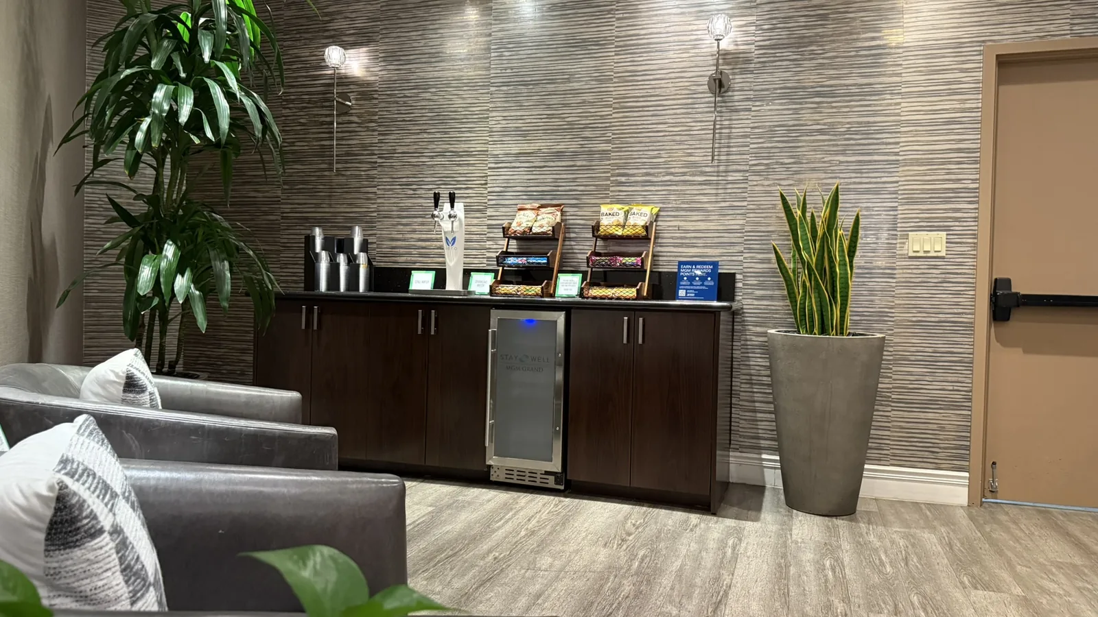 A quiet airport lounge with a self-serve coffee and snack station, comfortable armchairs, and plants