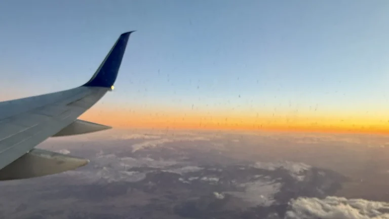 View from an airplane window of the wingtip against a sunset horizon over snow-covered mountains